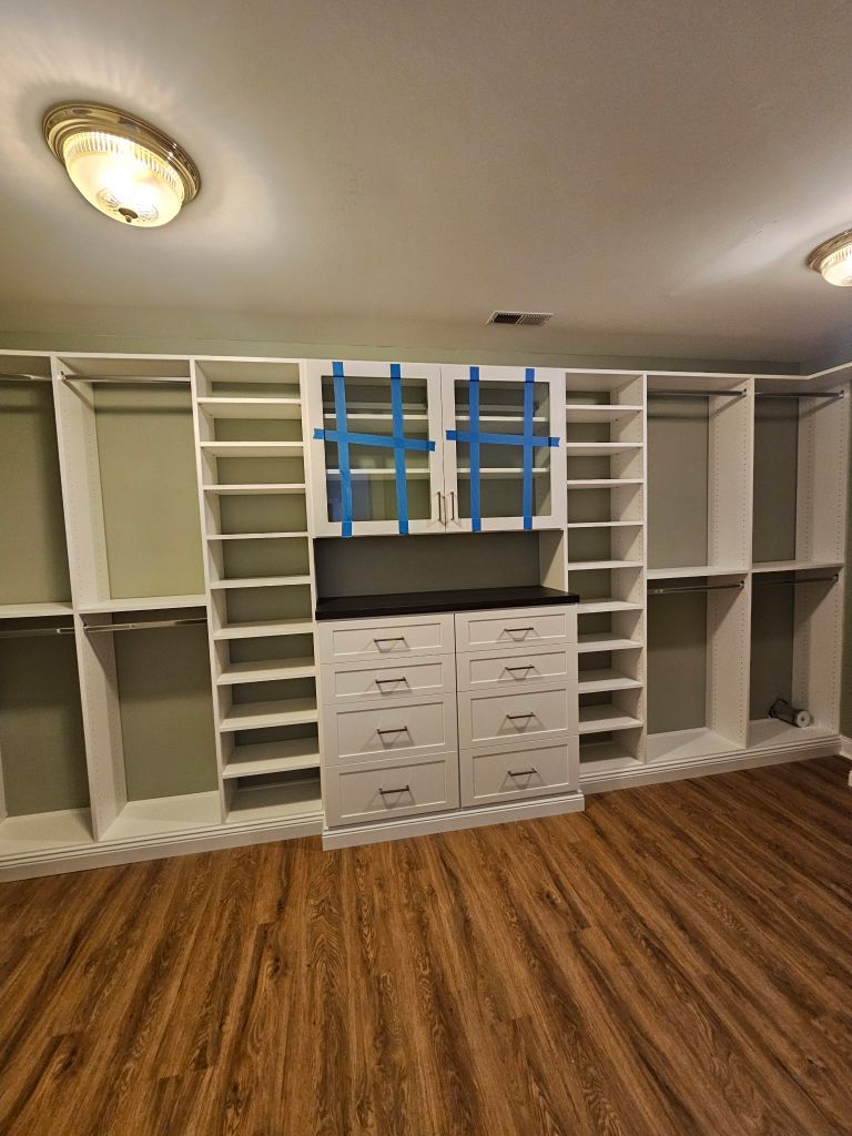 Empty white closet organizer with drawers and shelves on wooden floor.