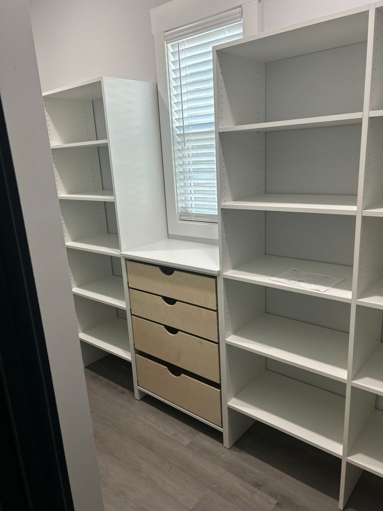 Organized walk-in closet with white shelves and wooden drawers under a window.